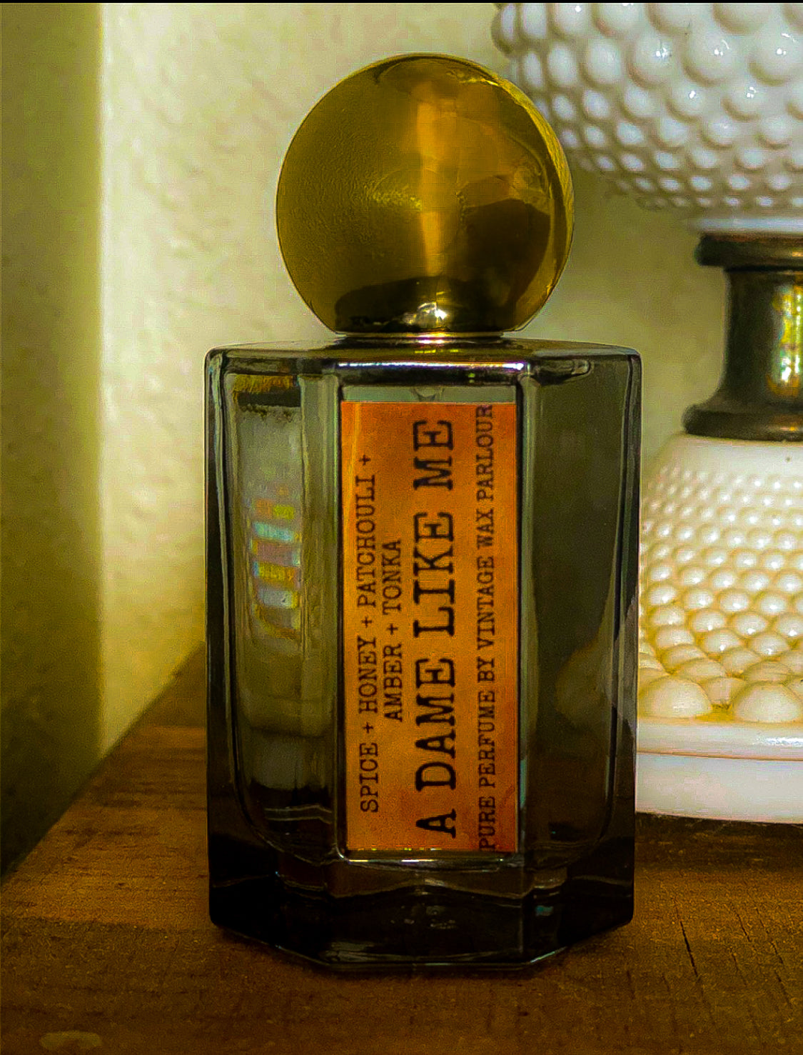 Perfume bottle labeled 'A Dame Like Me' on a wooden surface with a textured wall in the background.