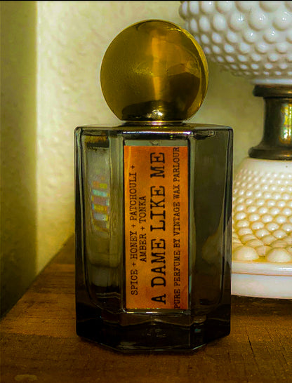 Perfume bottle labeled 'A Dame Like Me' on a wooden surface with a textured wall in the background.