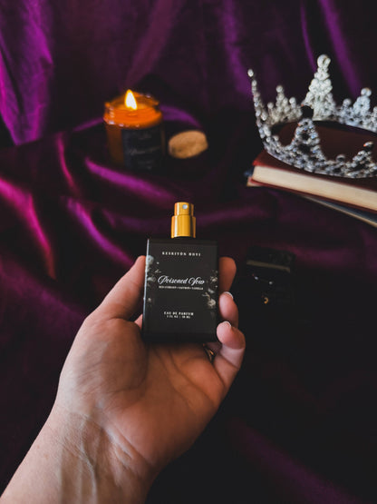 Hand holding a perfume bottle with a tiara and candle on a purple fabric background
